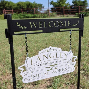 May include: Black metal welcome sign with the word "WELCOME" in green lettering and a decorative leaf design. A white sign below reads "LANGLEY METAL WORKS Established 2010" with an anvil graphic, hanging by chains.