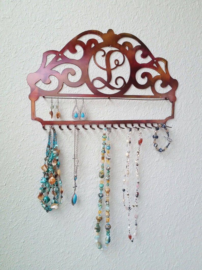 K&ouml;nnte beinhalten: Ein kupferfarbener Schmuckorganizer aus Metall mit einem dekorativen Schn&ouml;rkeldesign und dem Buchstaben "L" in der Mitte. Der Organizer hat Haken zum Aufh&auml;ngen von Halsketten und Ohrringen.