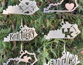 May include: Six silver metal ornaments shaped like the state of Kentucky. Each ornament features a different design, including a heart, the letters "K" and "Y", the word "Kentucky", the letters "UK", and the word "Kentucky".