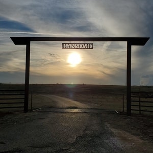 May include: A black metal gate with a sign that reads "RANSOME" hanging from it. The gate is open and leads to a dirt road. The sun is setting in the background, casting a warm glow over the scene.