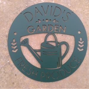 May include: A circular, dark teal metal sign with cut-out lettering and a watering can design. The text reads "DAVID'S GARDEN FRESH PRODUCE". Decorative elements include arrows, dots, and leaf sprigs. The sign is likely for a garden or farm.