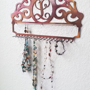 K&ouml;nnte beinhalten: Ein kupferfarbener Schmuckorganizer aus Metall mit einem dekorativen Schn&ouml;rkeldesign und einem einzigen Initial "L" in der Mitte. Der Organizer hat ein langes, rechteckiges Regal mit Haken darunter zum Aufh&auml;ngen von Halsketten und Ohrringen.
