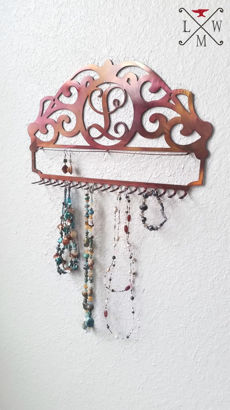 K&ouml;nnte beinhalten: Ein kupferfarbener Schmuckorganizer aus Metall mit einem dekorativen Schn&ouml;rkeldesign und einem einzigen Initial "L" in der Mitte. Der Organizer hat ein langes, rechteckiges Regal mit Haken darunter zum Aufh&auml;ngen von Halsketten und Ohrringen.