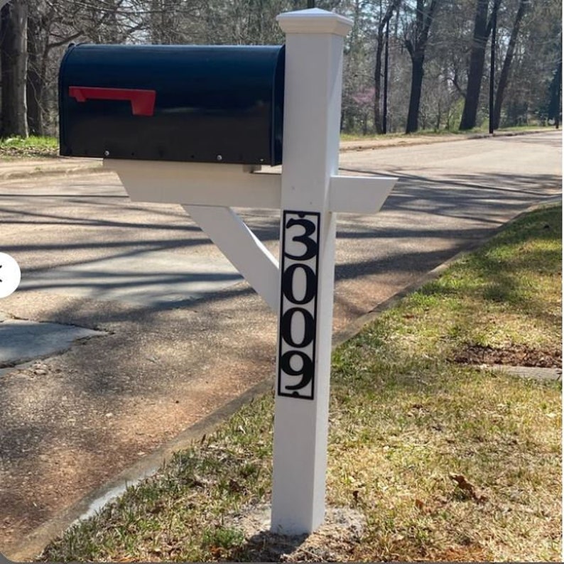 Op de afbeelding: Een witte brievenbuspaal met een zwarte brievenbus en een zwart metalen huisnummerplaatje met de tekst "3009".