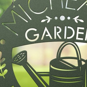 May include: Dark green metal garden sign with the word "GARDEN" in large, cut-out letters. The sign features a watering can and decorative leaf accents. The name "MICHEA" is above the word "GARDEN".
