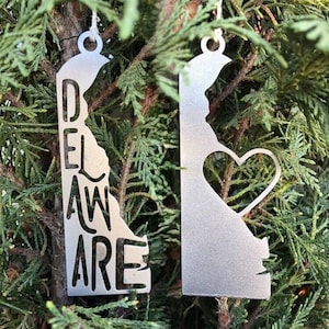 May include: Two silver metal ornaments shaped like the state of Delaware. One ornament has the word "DELAWARE" cut out of the state shape. The other ornament has a heart cut out of the state shape.