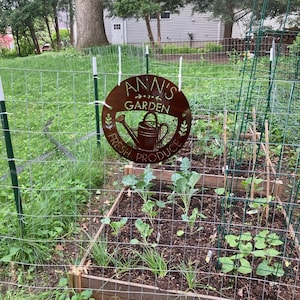 May include: A round, rusty-brown metal sign that reads "Ann's Garden Fresh Produce" with a watering can illustration. The sign hangs on a wire fence in front of a garden with raised beds and various plants.