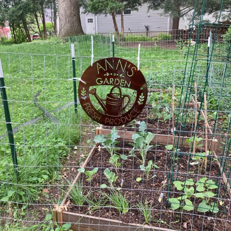 May include: A round, rusty-brown metal sign that reads "Ann's Garden Fresh Produce" with a watering can illustration. The sign hangs on a wire fence in front of a garden with raised beds and various plants.