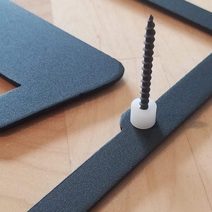 Op de afbeelding: Een zwart metalen frame met een schroef en een witte plastic dop. Het frame is bevestigd aan een houten oppervlak.