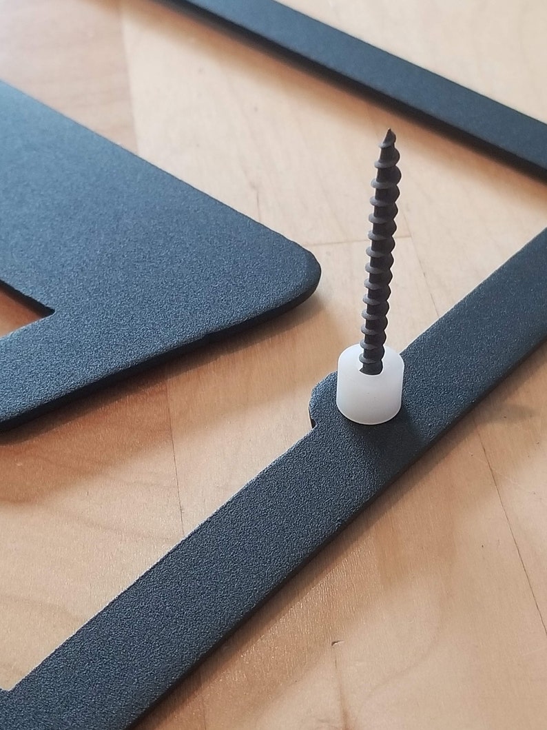 Op de afbeelding: Een zwart metalen frame met een schroef en een witte plastic dop. Het frame is bevestigd aan een houten oppervlak.