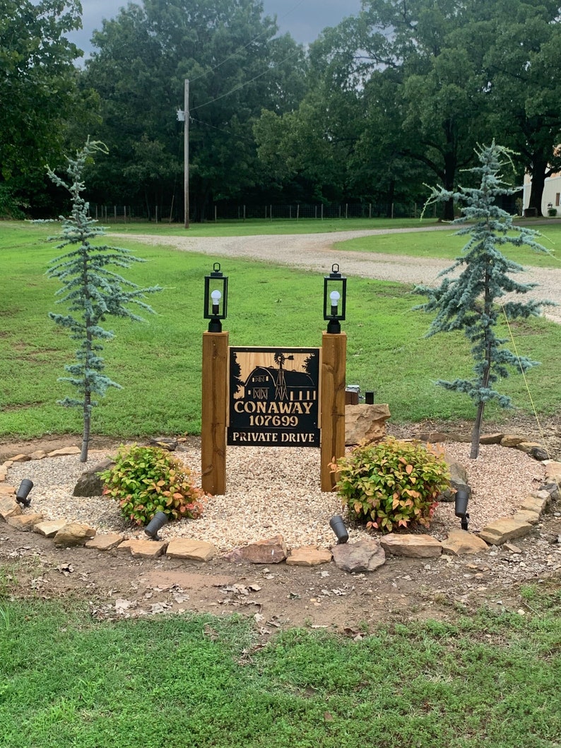 May include: A wooden sign with black lettering that reads "Hilltop Conway 107699 Private Drive" with two black lanterns on wooden posts on either side of the sign.