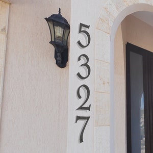 Pu&ograve; includere: Numeri civici in metallo nero 5327 montati su una parete bianca. I numeri sono in linea verticale e sono fissati con viti. Un apparecchio di illuminazione nero &egrave; montato sulla parete a sinistra dei numeri.
