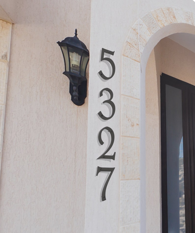Pu&ograve; includere: Numeri civici in metallo nero 5327 montati su una parete bianca. I numeri sono in linea verticale e sono fissati con viti. Un apparecchio di illuminazione nero &egrave; montato sulla parete a sinistra dei numeri.