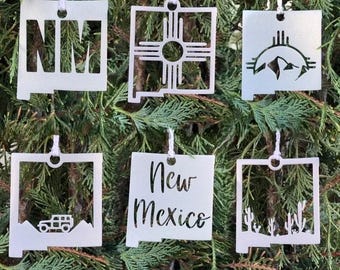May include: A collection of six silver-colored metal ornaments, each with a unique design. The ornaments are in the shape of the state of New Mexico, with cut-out designs including the letters "NM", the Zia symbol, mountains, a jeep, and cacti. The text "New Mexico" is also featured.