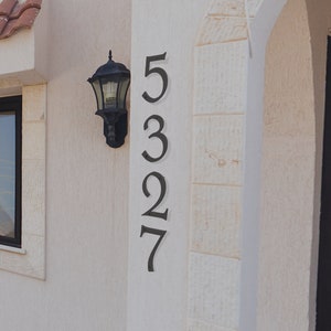Pu&ograve; includere: Un muro esterno bianco con una targa di numero civico in metallo nero. Il numero &egrave; 5327. Un apparecchio di illuminazione in metallo nero &egrave; montato sul muro.