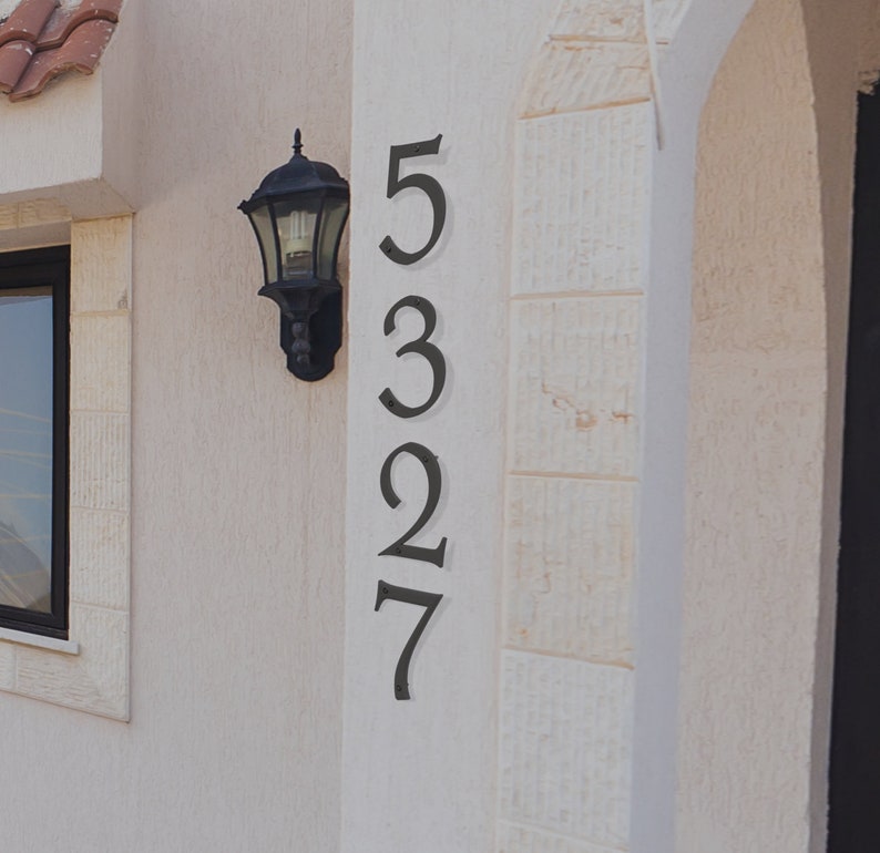Pu&ograve; includere: Un muro esterno bianco con una targa di numero civico in metallo nero. Il numero &egrave; 5327. Un apparecchio di illuminazione in metallo nero &egrave; montato sul muro.