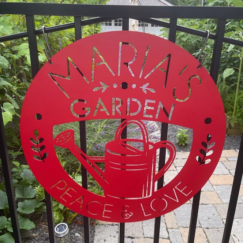 May include: A red metal garden sign with the words "MARIA'S GARDEN" and "PEACE & LOVE" cut out. A watering can is in the center. The sign is hanging on a black metal fence.