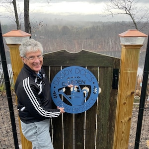 May include: A blue metal sign with the text "Daddy Doc's Garden Fresh Produce" and a watering can design. The sign is mounted on a wooden gate. The sign is held by a person wearing a black jacket with white stripes.