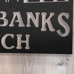 May include: A black metal sign with white lettering that reads "The Banks Ranch".