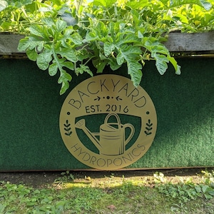 May include: A round, gold-colored sign with the words "BACKYARD HYDROPNICS EST. 2016" in a circular design. The sign features a watering can illustration. The sign is mounted on a green surface with green leafy plants growing above.
