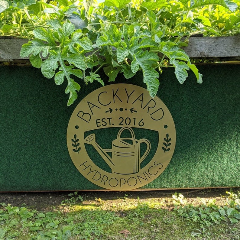 May include: A round, gold-colored sign with the words "BACKYARD HYDROPNICS EST. 2016" in a circular design. The sign features a watering can illustration. The sign is mounted on a green surface with green leafy plants growing above.