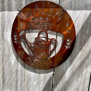 May include: A round, rust-colored metal sign with the words "WALSH STREET GARDEN" cut out. A watering can is in the center. The sign is hanging on a weathered wooden fence.