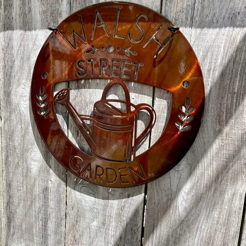 May include: A round, rust-colored metal sign with the words "WALSH STREET GARDEN" cut out. A watering can is in the center. The sign is hanging on a weathered wooden fence.