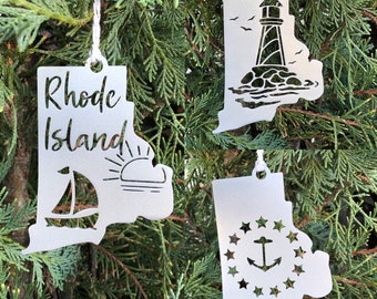 May include: Three metal ornaments shaped like the state of Rhode Island. The first ornament features the text "Rhode Island" and a sailboat and sun. The second ornament features a lighthouse. The third ornament features the state seal with an anchor and stars.