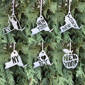 May include: Six silver metal ornaments shaped like the state of New York. The ornaments feature different designs, including the words "New York", "I Love NY", and an apple with the words "New York".
