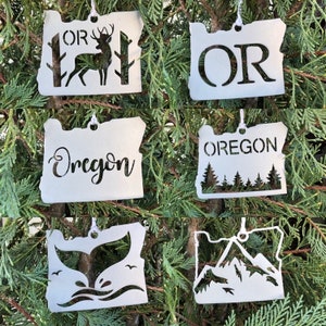 May include: Six white metal ornaments shaped like the state of Oregon, each with a different design. The designs include a deer, the letters "OR", the word "Oregon", a whale tail, a mountain range, and a group of pine trees.