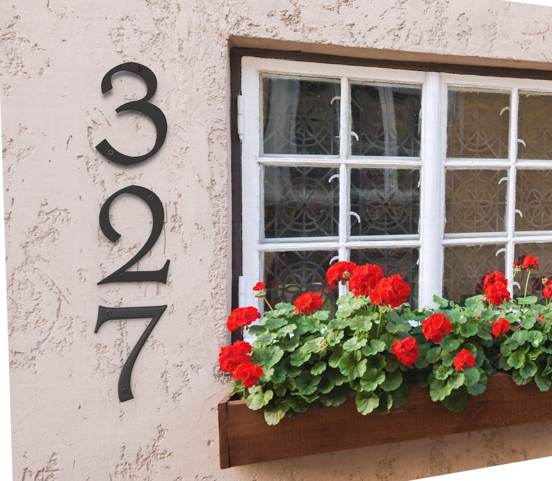 Pu&ograve; includere: Numeri civici in metallo nero 327 montati su un muro in stucco beige accanto a una finestra con cornice bianca. Gerani rossi piantati in una fioriera di legno.