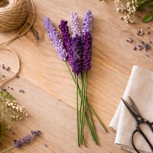 Op de afbeelding: Een bundel handgemaakte lavendeltakken in verschillende tinten paars, van licht lavendel tot diep violet. De stelen zijn groen en de bloemen zijn gerangschikt op een licht houten oppervlak. Een bol touw, een schaar en een linnen doek zijn ook aanwezig.