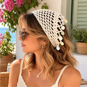 May include: A cream-colored crochet headscarf featuring an open diamond-shaped mesh pattern and a decorative border adorned with small, dangling circular metallic discs that catch the light, creating a textured and layered appearance.