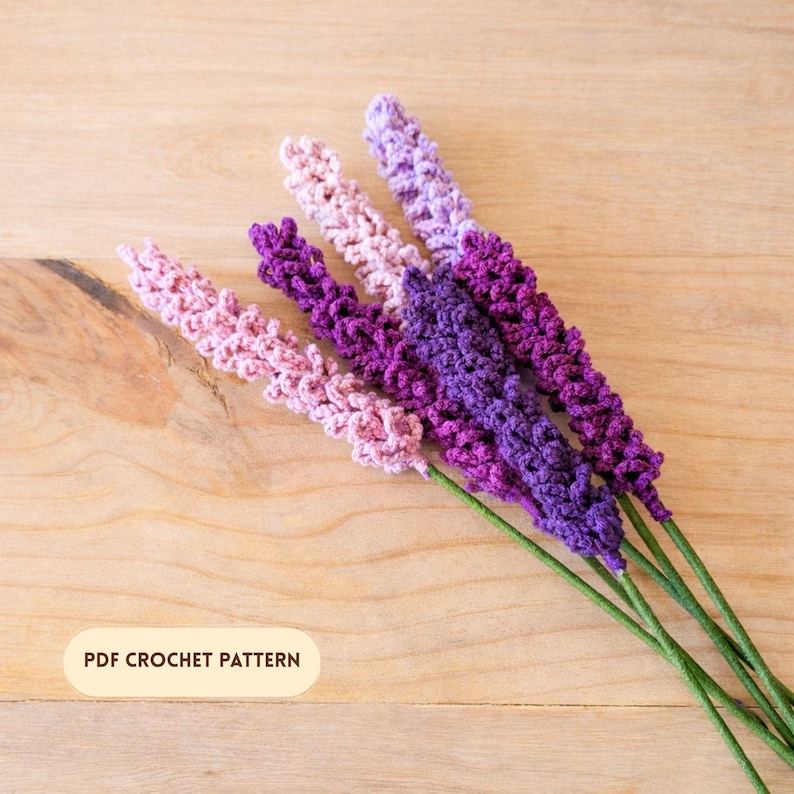 Op de afbeelding: Een verzameling handgemaakte gehaakte lavendelstelen in roze, paarse en lavendel tinten. De stelen hebben groene stelen en zijn gerangschikt op een licht houten oppervlak. De tekst "PDF CROCHET PATTERN" is zichtbaar.