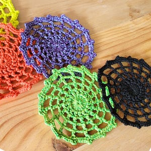 May include: A collection of colorful, handmade crochet coasters. The coasters are in shades of orange, purple, green, yellow, and black. Each coaster has a circular, openwork design, and they are arranged on a wooden surface.