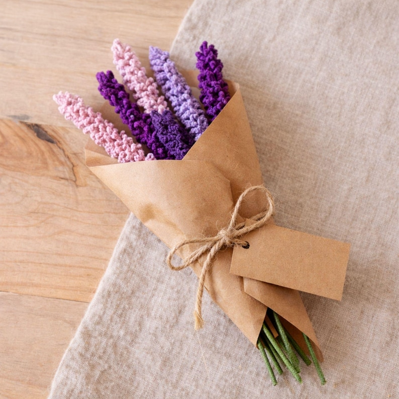 Op de afbeelding: Een boeket handgemaakte gehaakte lavendelbloemen in roze, paarse en violette tinten, verpakt in bruin papier. Het boeket is vastgebonden met touw en bevat een blanco label. De stelen zijn groen.