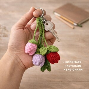 May include: A handmade crochet keychain with three flower buds in pink, purple, and red. The buds are attached to a green stem with green leaves. A silver key ring and a key are also attached. The text on the right says: Bookmark, Keychain, Bag Charm.