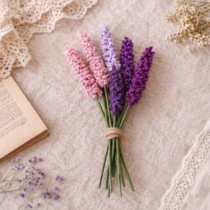 Op de afbeelding: Een boeket handgemaakte gehaakte lavendelbloemen in tinten roze, paars en violet, vastgebonden met touw. De stelen zijn groen. De bloemen zijn gerangschikt op een neutraal gekleurd linnen oppervlak.