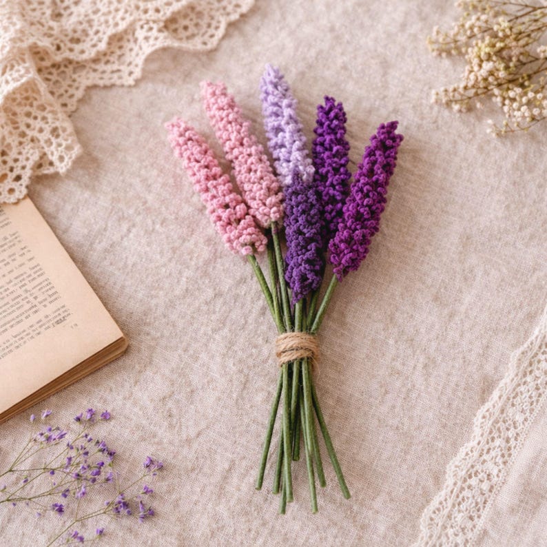 Op de afbeelding: Een boeket handgemaakte gehaakte lavendelbloemen in tinten roze, paars en violet, vastgebonden met touw. De stelen zijn groen. De bloemen zijn gerangschikt op een neutraal gekleurd linnen oppervlak.