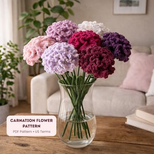 Op de afbeelding: Een boeket handgemaakte gehaakte anjerbloemen in een heldere glazen vaas. De bloemen zijn in tinten roze, paars, rood en wit. De vaas staat op een houten tafel. De tekst "CARNATION FLOWER PATTERN" is zichtbaar.
