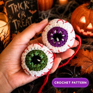 May include: Two round crochet eyeballs featuring white yarn with red embroidered veins. One eyeball has a purple iris and the other has a green iris, both with black centres. A red yarn string connects the two pieces, and a purple label reads CROCHET PATTERN.