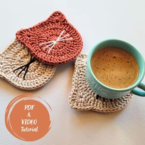 May include: Three handmade crochet cat-shaped coasters in shades of tan and rust-orange. One coaster holds a teal mug filled with coffee. The coasters have embroidered details for the cat's face. The image also includes text that says "PDF & VIDEO Tutorial".