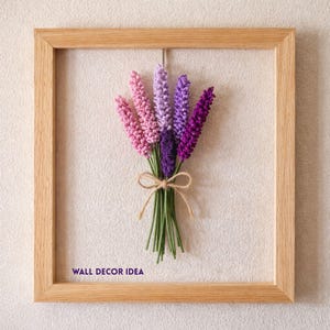 Op de afbeelding: Een ingelijste wanddecoratie met een boeket lavendelbloemen in roze, paarse en violette tinten. De bloemen zijn vastgebonden met touw en geplaatst tegen een neutrale achtergrond. De houten lijst geeft een rustieke uitstraling. Tekst linksonder: WALL DECOR IDEA.