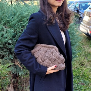 May include: A brown knitted clutch bag with a woven design. The bag is held by a person wearing a navy coat. The clutch is a neutral colour and appears to be made of soft yarn.