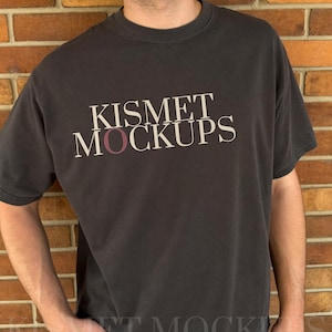 May include: A charcoal grey short-sleeve t-shirt featuring the text KISMET MOCKUPS printed in a serif font across the chest. The letter O in the word MOCKUPS is highlighted in a muted maroon colour, while the remaining letters are cream.