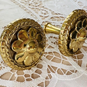 May include: Two ornate, gold-colored curtain tiebacks. Each tieback features a floral design in the center, surrounded by a decorative border. The hardware is a bright gold color, and the tiebacks are set against a white lace tablecloth.