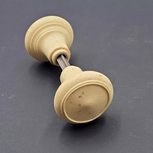 Puede incluir: Pomo de puerta vintage de color crema con una varilla metálica. El pomo tiene forma redondeada con una base ligeramente ovalada. El herraje muestra signos de edad y desgaste, con algo de decoloración y pequeñas manchas oscuras.