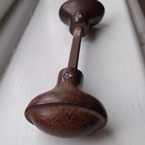 Puede incluir: Un pomo de puerta marrón vintage con forma redondeada y varilla de conexión rectangular. El pomo tiene un aspecto envejecido y desgastado, lo que sugiere que es antiguo. La ferretería es probablemente de metal.