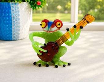 Könnte beinhalten: Eine Glasfigur eines hellgrünen Frosches mit großen roten und gelben Augen, der eine durchscheinende bernsteinfarbene Gitarre spielt. Der Frosch hat kleine schwarze Punkte an den Füßen sowie einen blauen und weißen Fleck auf dem Kopf.