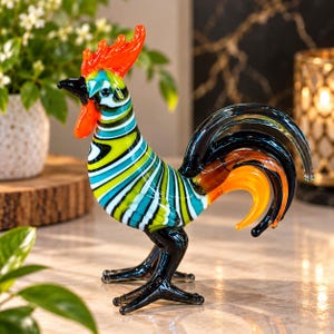 Puede incluir: Una figura de gallo de cristal con un vibrante patrón de rayas en tonos verde lima, azul cerceta y blanco. La escultura tiene una cresta y barbillas de color naranja brillante, un pico negro, patas negras y una cola curva con detalles de cristal negro y naranja.